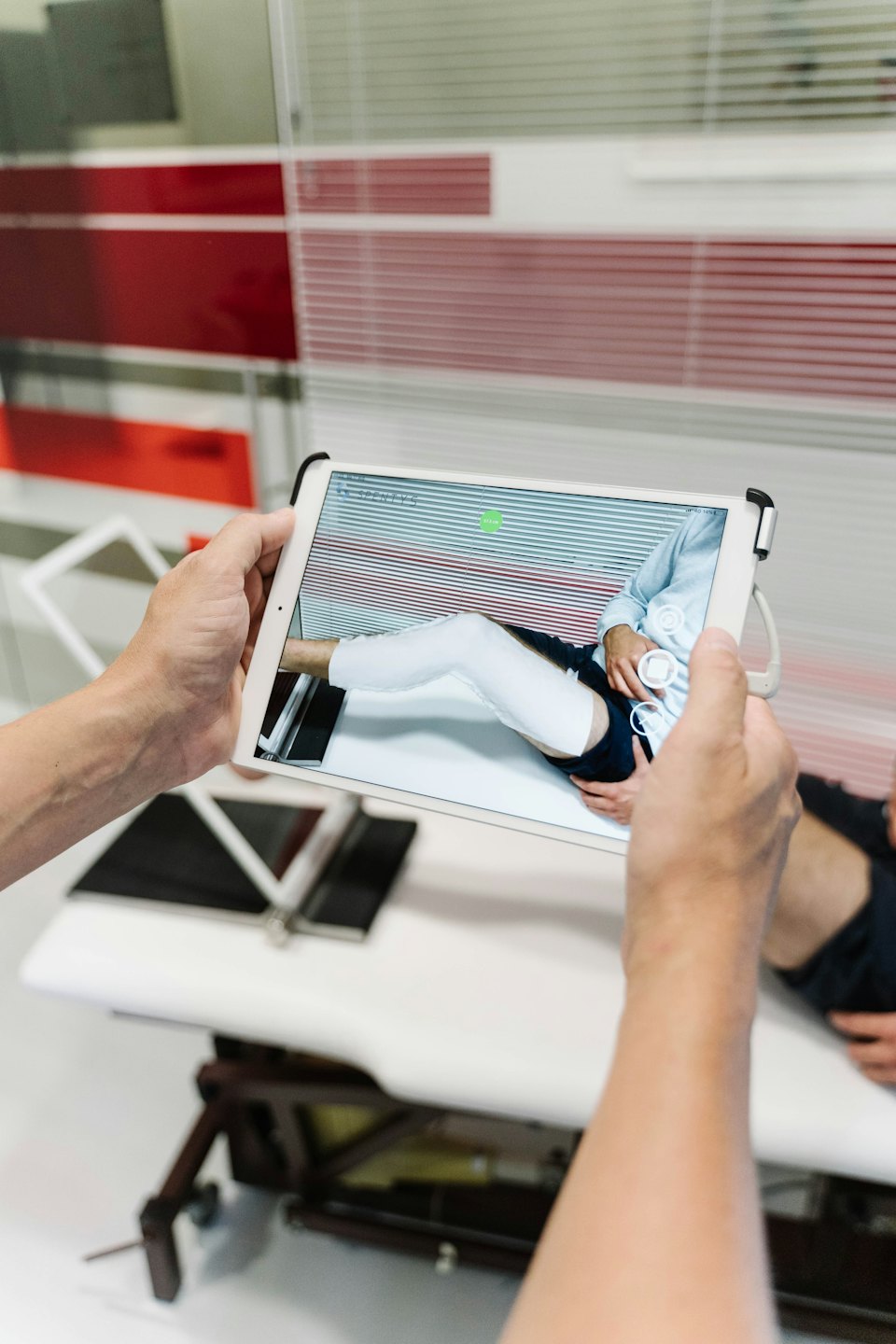 Hands holding tablet showing patient leg on examination table.
