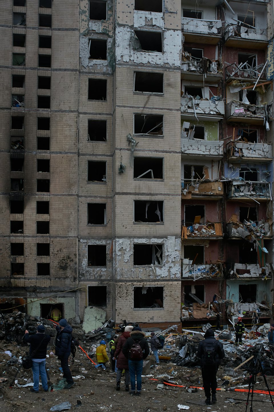 People stand among rubble in front of a damaged apartment building.